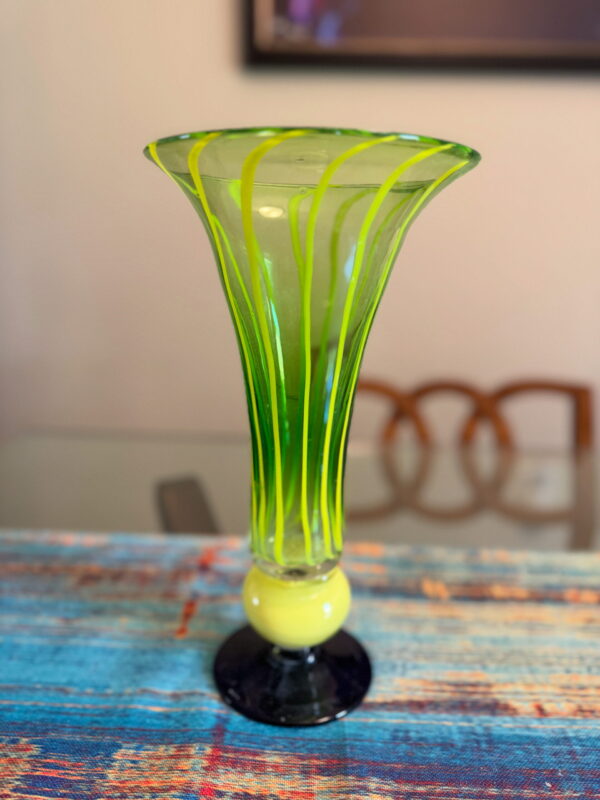 A glass vase with a flared rim and green and yellow vertical stripes stands on a dark blue base. It is placed on a colorful, textured blue surface with wooden chairs and a wall art piece blurred in the background.