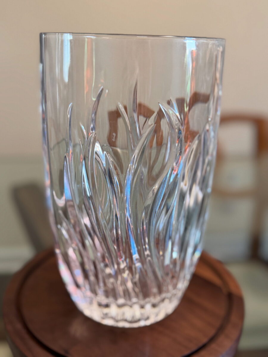 A clear glass vase with swirling, flame-like patterns is placed on a round wooden surface. The glass design creates dynamic, textured reflections. The background is softly blurred, with hints of furniture and neutral-colored walls.