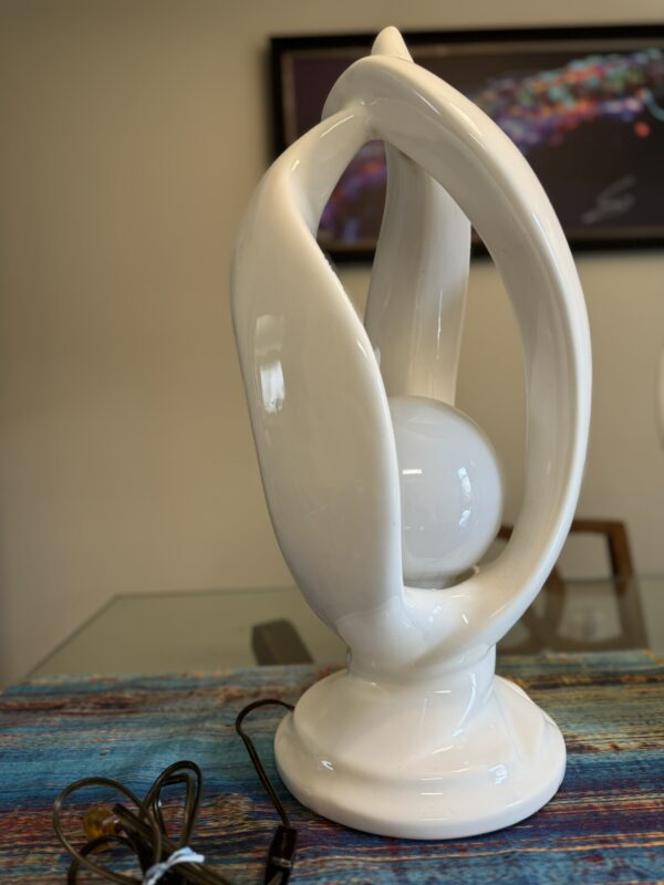 A glossy, modern white ceramic lamp with curved, leaf-like shapes forms an open structure around a central sphere. The lamp sits on a circular base on a colorful table, with its black cord visible. A blurred abstract painting hangs on the wall in the background.