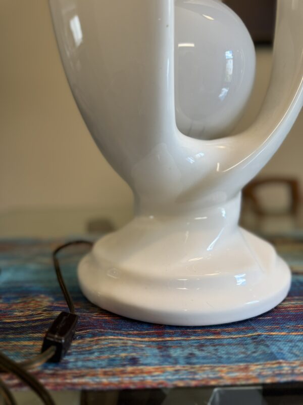 A glossy white ceramic lamp base with a modern, abstract design sits on a colorful woven table runner. A black power cord with a USB connector lies on the glass tabletop beside the lamp. The background is softly blurred.