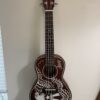 A dark brown Lanikai ukulele with silver tuning pegs and white decorative artwork of an eagle and stars on the body, hangs on a light-colored wall near a window with white blinds.