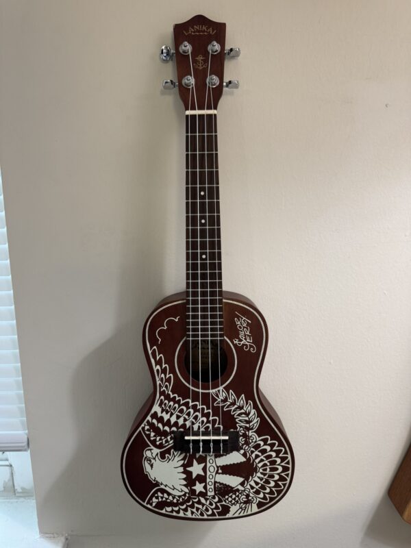 A dark brown Lanikai ukulele with silver tuning pegs and white decorative artwork of an eagle and stars on the body, hangs on a light-colored wall near a window with white blinds.