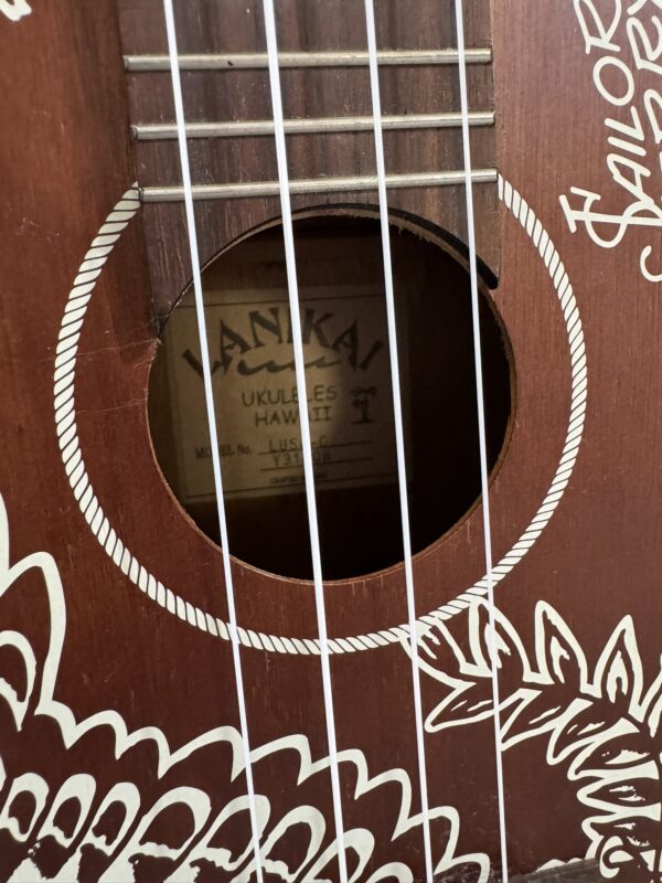 Close-up of a wooden ukulele’s sound hole and four white strings. The body features intricate white floral and geometric designs. Inside the sound hole, a label reads “Lanikai Ukuleles Hawaii.” The wood grain and details of the instrument are visible.