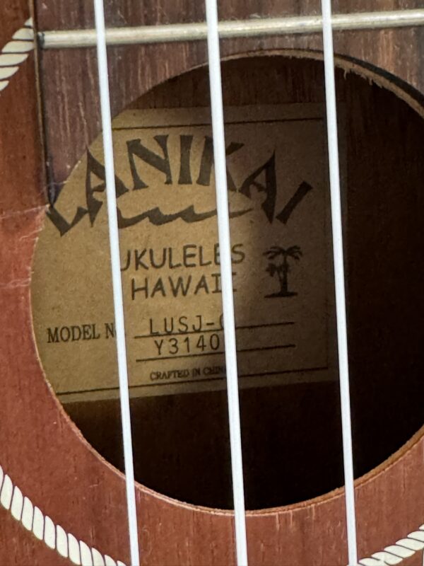 Close-up of a ukulele’s sound hole showing a label inside. The label reads Lanikai Ukuleles Hawaii with a palm tree graphic, model number LUSJ-Y3140, and Crafted in China. Three white strings cross in front of the label.