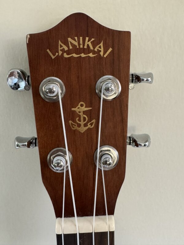 Close-up of a wooden ukulele headstock with silver tuning pegs. The brand name LANIKAI and a gold anchor symbol are printed on the wood. Two white strings are visible, and the background is a plain, light-colored wall.