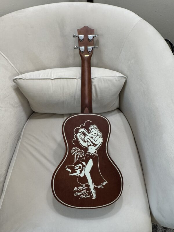 A wooden ukulele rests on a beige armchair. The back of the ukulele is decorated with a white illustration of a hula dancer, a palm tree, and the text “ALOHA HAWAII 1982.” The design stands out against the dark wood.