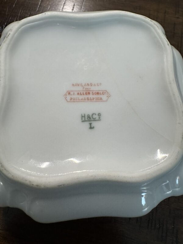 A close-up of the bottom of a white porcelain dish with a scalloped edge. Stamped in red is HAVILAND & Co., for R.J. ALLEN & SONS, PHILADELPHIA. Below, in green, is H&C° L. The dish rests on a wooden surface.