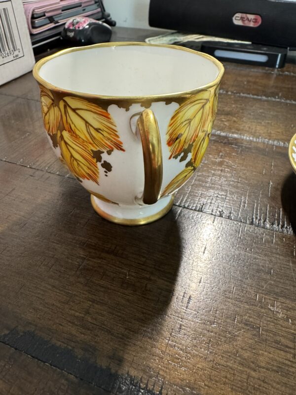 A porcelain teacup with gold trim and handle sits on a wooden table. The cup is decorated with yellow and orange leaf patterns and has some worn gold detailing. In the background, there are office supplies, a paper box, and a black electronic device.