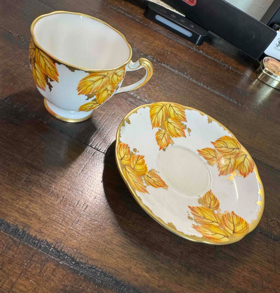 A porcelain teacup and saucer with gold handles and rims sit on a wooden table. Both pieces feature yellow and gold autumn leaves in a delicate pattern. The saucer is to the right of the cup, set at an angle.