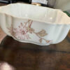 A white, scalloped-edge ceramic bowl with pale pink and brown floral designs sits on a wooden table. The bowl’s surface has slight imperfections, and the background includes blurred office supplies and a partially visible plastic bag.