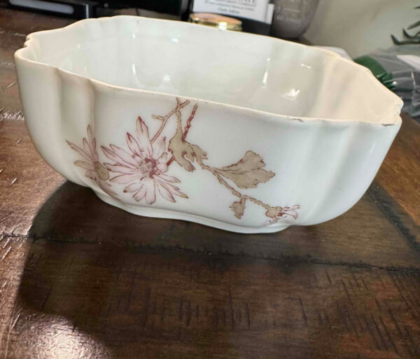 A white, scalloped-edge ceramic bowl with pale pink and brown floral designs sits on a wooden table. The bowl’s surface has slight imperfections, and the background includes blurred office supplies and a partially visible plastic bag.