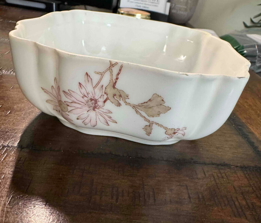 A white, scalloped-edge ceramic bowl with pale pink and brown floral designs sits on a wooden table. The bowl’s surface has slight imperfections, and the background includes blurred office supplies and a partially visible plastic bag.
