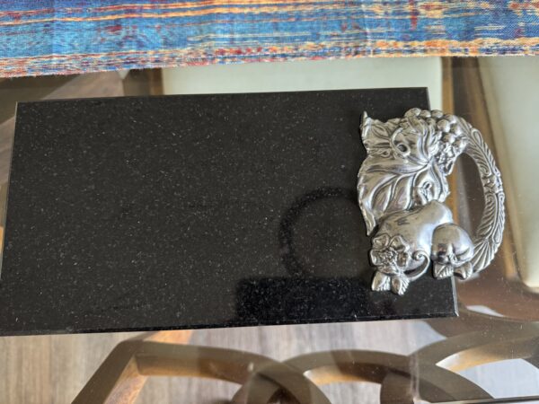 A black rectangular stone tray sits on a glass table. The right side features an ornate silver handle shaped like a cornucopia, overflowing with fruit and leaves. Colorful woven fabric appears at the top edge of the image.