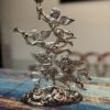 A silver-colored metal sculpture of four flying cherubs with wings is shown. One cherub holds a torch aloft, another plays a trumpet, and two play stringed instruments. The sculpture rests on a cloud-like base, and is set on a colorful, patterned tablecloth.