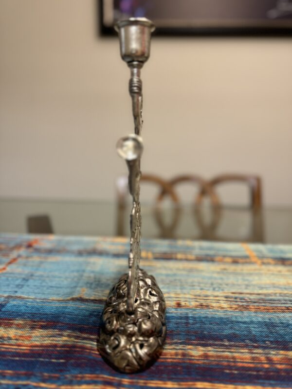 A decorative silver candle holder with an ornate, textured base and a single candle cup stands on a colorful, woven table runner. In the background, there are three wooden chairs and a blurred framed picture on a beige wall.