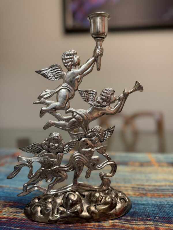 A silver-toned metal figurine of three winged cherubs in flight. The top cherub holds a candle holder, the middle one blows a horn, and the bottom cherub plays a harp; all are attached to an ornate base on a colorful woven tablecloth.