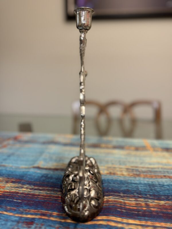 A silver candlestick holder with an ornate, floral design sits on a colorful woven tablecloth. The holder is viewed from the narrow end, with a glass table and two blurred wooden chairs in the background. The scene is softly lit.