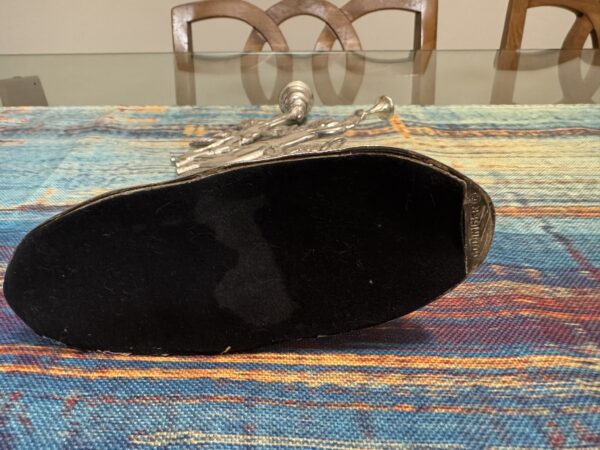 A silver figurine lies on its side on a colorful, patterned tablecloth atop a glass table. The focus is on the black-felted base of the figurine, with a small metallic label that reads Godinger. Wooden chairs are visible in the background.