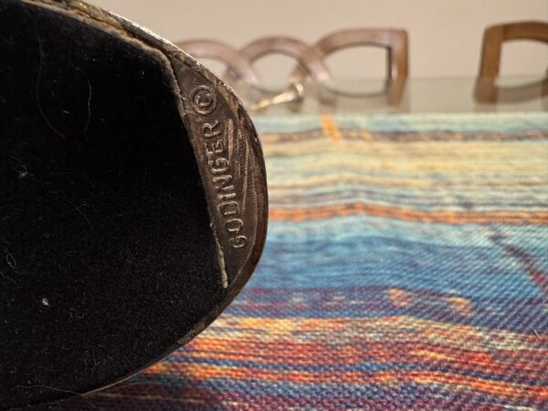 Close-up of a metal object engraved with GODINGER resting on a colorful woven fabric. In the background, four wooden chairs and the edge of a glass table are visible, slightly out of focus.