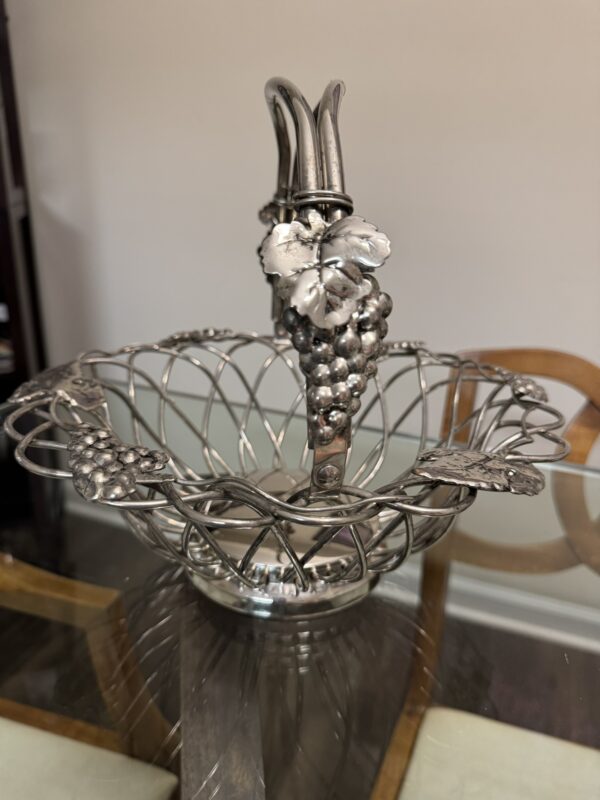 A decorative silver metal fruit basket with a woven openwork design and vine-shaped handle, adorned with detailed grape clusters and leaves. The basket sits on a glass table, with parts of wooden chairs visible in the background.