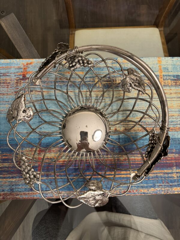 A round, silver metal wire fruit basket with grape and leaf decorations sits on a colorful, weathered wooden table. The basket has an intricate spiral pattern and a reflective center showing the photographer’s reflection. A chair is partially visible behind the table.