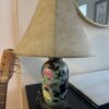 A decorative table lamp with a beige, textured lampshade sits on a black surface. The lamp base is black with colorful floral patterns. Part of an acoustic guitar and a closed laptop are visible nearby, with a round mirror on the wall in the background.