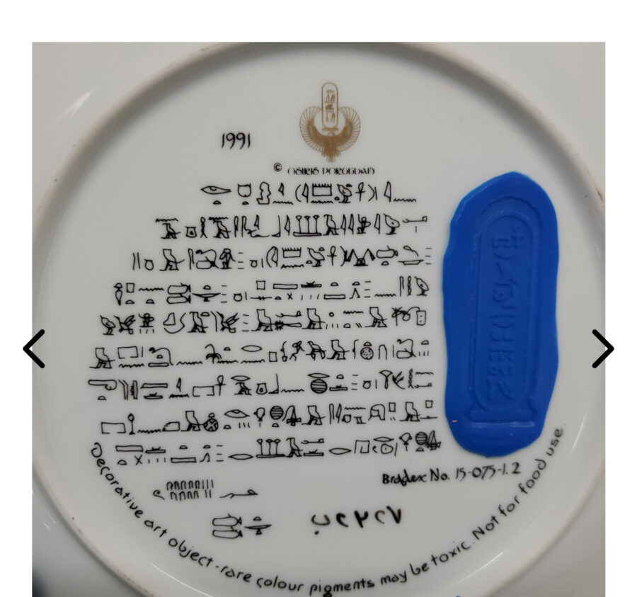 A white plate features black Egyptian hieroglyphs and a blue cartouche on the right. Above the hieroglyphs are the year 1991 and a gold emblem. Below is English text about rare pigments being toxic and a note: Not for food use. Arrows flank the image.