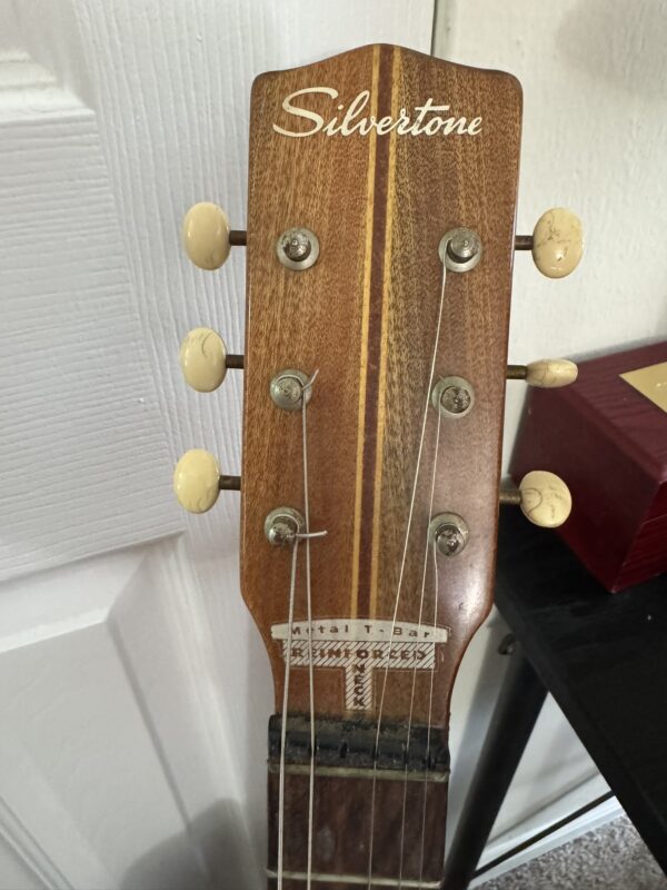 Close-up of a vintage Silvertone guitar headstock, showing six tuning pegs with cream-colored knobs. Two strings appear to be missing. The headstock has a wood grain finish, Silvertone logo at the top, and part of the nut and fretboard are visible.