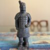 A small, detailed statue of a Terracotta Warrior stands on a colorful, patterned surface. The warrior wears traditional armor and has one hand clenched, the other relaxed. Out-of-focus chairs and a framed picture appear in the background.
