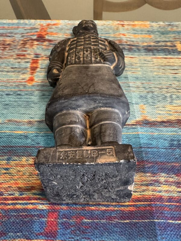 A small black terracotta warrior figurine lies on its back on a colorful, textured fabric. The warrior wears detailed armor and has Chinese characters inscribed on the rectangular base near its feet. The image is taken from a low angle.