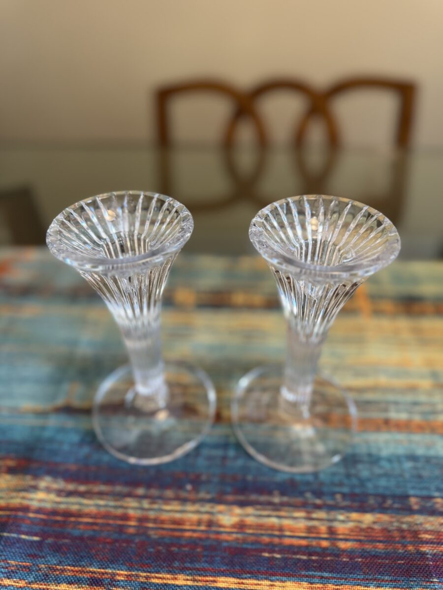 Two clear, fluted glass vases with wide, scalloped rims stand side by side on a colorful, woven table runner. In the background, a blurred wooden chair and a glass table are visible. The vases have a decorative, ribbed texture.