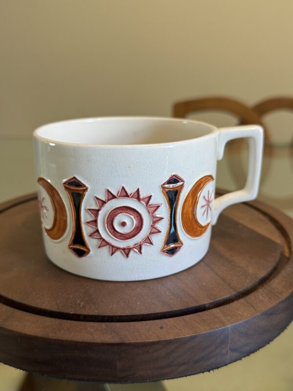 A white ceramic mug with a square handle sits on a round wooden tray. The mug features a colorful geometric pattern of red and orange suns, moons, and black diamonds in relief, arranged in a repeating design around its side. The background is blurred.