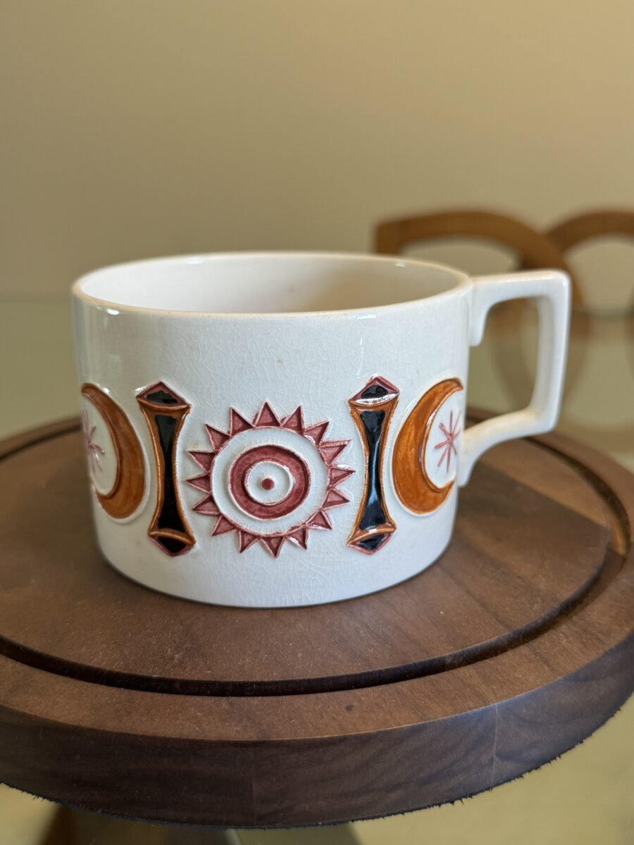A white ceramic mug with a square handle sits on a round wooden tray. The mug features a colorful geometric pattern of red and orange suns, moons, and black diamonds in relief, arranged in a repeating design around its side. The background is blurred.
