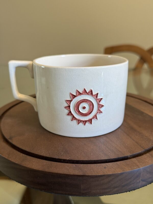 A white ceramic mug with a square handle sits on a round wooden tray. The mug features a red sun and eye design embossed on its side. The background is softly blurred, showing part of a wooden chair and a beige wall.