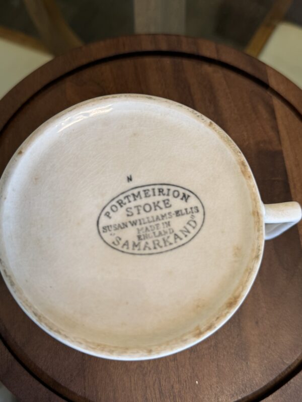 A close-up of the underside of a white teacup shows black printed text reading “PORTMEIRION STOKE SUSAN WILLIAMS-ELLIS MADE IN ENGLAND SAMARKAND,” surrounded by an oval border. The teacup sits on a round wooden surface.
