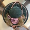 A plush, round green velvet hat seen from above, adorned with a dusty pink fabric rose, green-brown ribbon leaves, and pink twisted cord trim. The hat sits on a glass table with cream and wooden chairs visible in the background.
