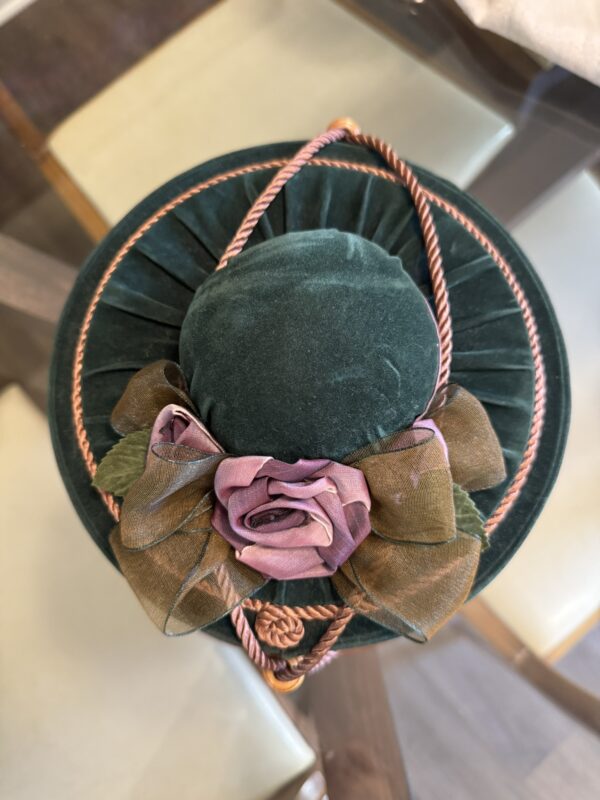 A plush, round green velvet hat seen from above, adorned with a dusty pink fabric rose, green-brown ribbon leaves, and pink twisted cord trim. The hat sits on a glass table with cream and wooden chairs visible in the background.