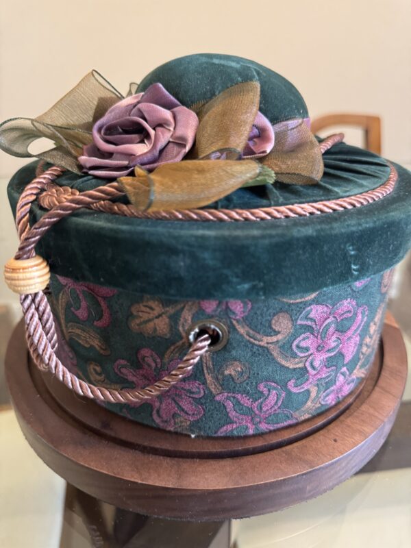 A round, green velvet box with purple floral patterns and a matching lid. The lid is decorated with purple and gold ribbon flowers and leaves. A twisted gold and purple cord is looped around the box, which rests on a wooden base.