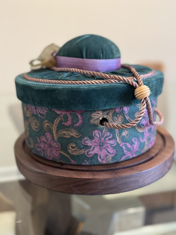 A round, decorative box with a green velvet lid and purple trim, adorned with floral patterns in gold and purple. A twisted cord with a wooden bead is draped over the box, which sits on a wooden base. The background is softly blurred.