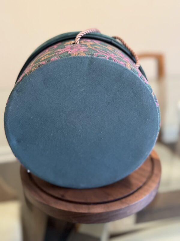 A round, teal-colored box with a soft fabric bottom rests on its side atop a wooden surface. The box has a floral-patterned exterior and a rope handle, while the background shows blurred furniture.