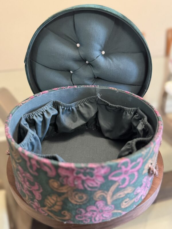 A round, fabric-covered box with a tufted, cushioned lid stands open. The inside is lined with dark fabric and features gathered pockets along the sides. The exterior is decorated with a pink and gold floral pattern.