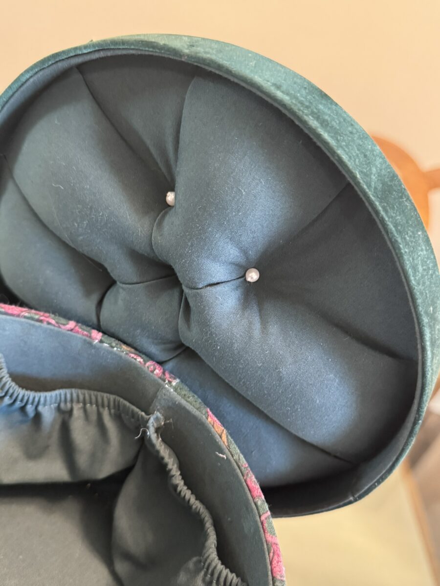 A close-up of an open, round, green velvet box with a fabric-lined lid. The lid features tufted dark fabric with two small, round, pearl-like buttons in the center. The box’s interior edge is trimmed with a colorful, patterned fabric.