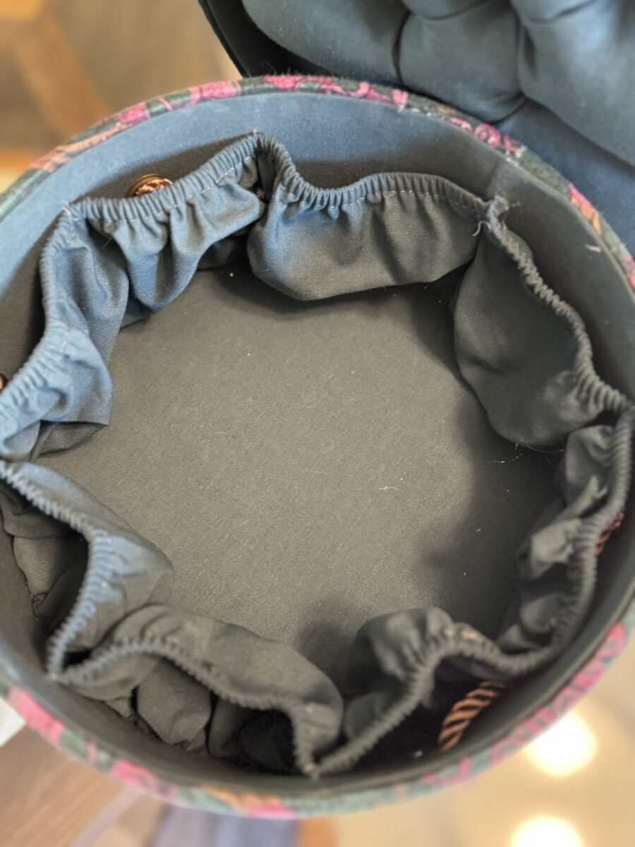 A round, fabric-lined container with gathered, elastic black pockets along the inner sides. The main compartment is empty, and the fabric has a dark, solid color. The rim has a bit of pinkish pattern visible around the edge.