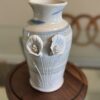 A handmade ceramic vase with a glossy cream and blue glaze sits on a round wooden stand. The vase features raised floral decorations with two sculpted leaves and small white flower buds on its front. The background is softly blurred.