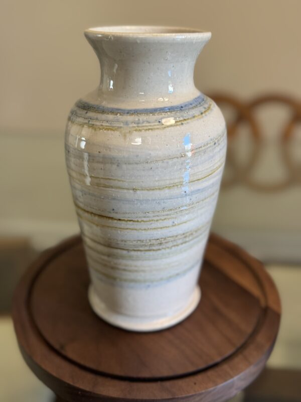 A tall, glossy ceramic vase with a wide rim and narrow base sits on a round wooden stand. The vase features horizontal, earthy-toned stripes in cream, blue, and tan. The blurred background shows part of a circular wooden object or chair.