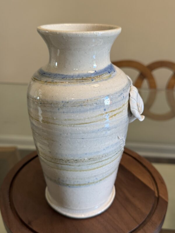 A ceramic vase with a glossy, off-white glaze featuring pale blue and tan horizontal streaks sits on a round wooden base. The vase has a flared lip and a small decorative white flower attached to its neck. A glass table and two wooden chairs are in the background.