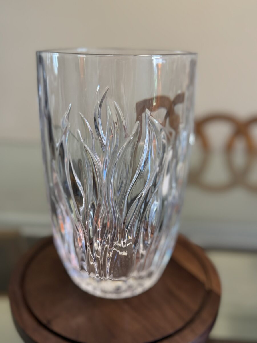 A clear glass vase with swirling, flame-like textured patterns stands on a round wooden surface. The background is softly blurred, showing part of a light wall and a decorative brown circular object. The vase catches and reflects light.