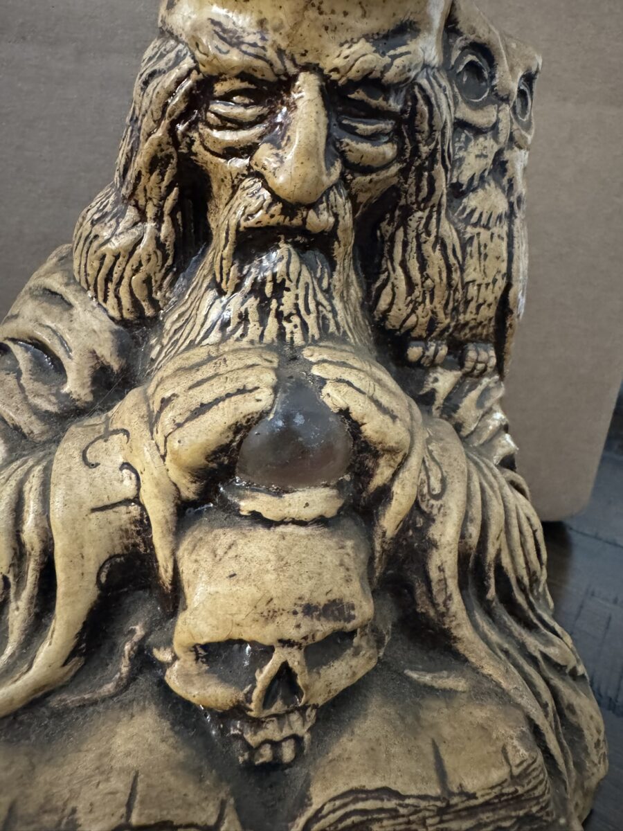 A close-up of a detailed, brownish stone or ceramic statue depicting a bearded wizard holding a crystal ball above a skull, with an owl perched on his shoulder. The statue is textured and intricately carved, set against a plain background.