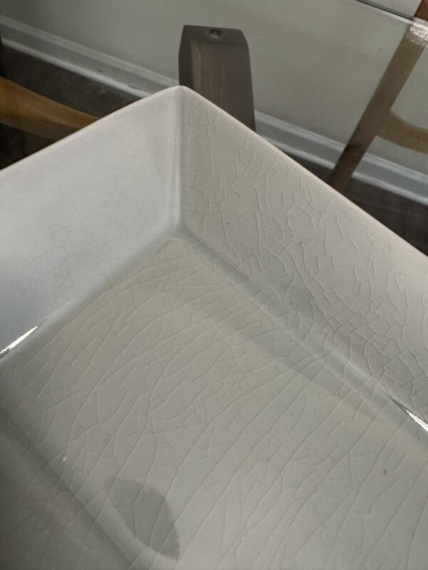 A close-up of a white, square ceramic dish with visible crackle patterns in the glaze. The dish contains a shallow layer of clear water. In the background, parts of a glass table, a wall, and wooden chair legs are partially visible.