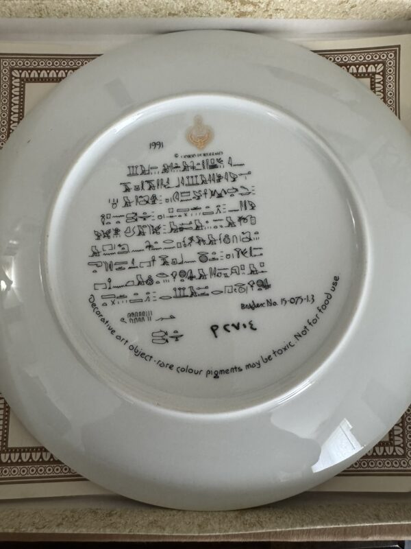 A decorative white plate features black printed Egyptian hieroglyphics and English text on its back. The English text warns that rare color pigments may be toxic and not for food use. Theres a logo and the year 1991 printed at the top.